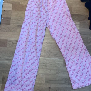 Rosa Barbie-byxor pyjamas  - Säljer ett par rosa byxor med ett all-over Barbie-tryck. Byxorna har en loose passform och är perfekta för en avslappnad stil. De är elastiska i midjan för extra komfort.