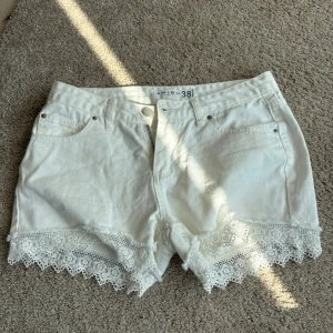 Vita jeansshorts med spets  - Säljer ett par vita jeansshorts med snygga spetsdetaljer längs bensluten. Shortsen har klassisk femficksdesign, knapp och dragkedja framtill samt normal passform. Perfekta för sommardagar!