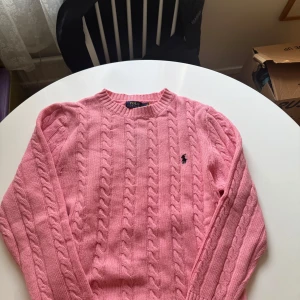 Rosa kabelstickad tröja från Ralph Lauren - Säljer en klassisk rosa kabelstickad tröja från Ralph Lauren med rund halsringning och broderad logga på bröstet. Tröjan har långa ärmar och är perfekt för lager-på-lager-stil.