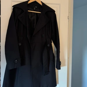 Svart trenchcoat från H&M - Snygg svart trenchcoat från H&M med klassisk design. Jackan har knappar framtill och ett bälte i midjan för en stilren look. Perfekt för våren med sina långa ärmar och tidlösa stil.