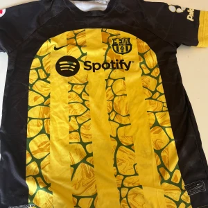 Gul och svart FC Barcelona fotbollströja - Säljer en unik FC Barcelona fotbollströja i gult och svart med ett iögonfallande mönster. Tröjan har korta ärmar och är prydd med sponsorloggor samt spelarens namn och nummer på ryggen. Perfekt för fans som vill sticka ut!