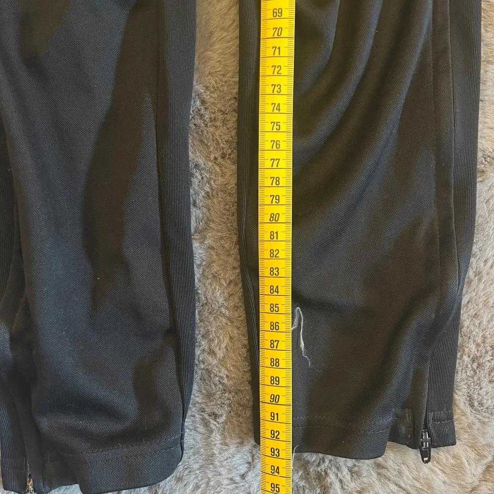 Snygga svarta träningsbyxor från Adidas med de klassiska tre vita ränderna längs sidorna. Byxorna har en smal passform och dragkedjor vid bensluten för enkel på- och avtagning. Perfekta för träning.. Farkut & Housut.