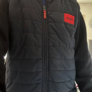 Svart quiltad jacka från Hugo - Snygg svart quiltad jacka från Hugo med dragkedja och röd logotypdetalj på bröstet. Perfekt för lager-på-lager under kyliga dagar. Västen har en stilren design med praktiska fickor.