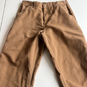 Bruna cargobyxor från Carhartt - Säljer ett par bruna cargobyxor från Carhartt. Byxorna har flera praktiska fickor och en knapp med Carhartt-logga. Perfekta för en avslappnad stil med en robust känsla.