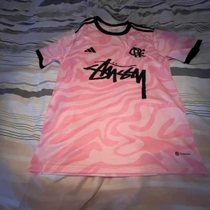 Rosa Stüssy x Adidas fotbollströja M - Säljer en limited edition fotbollströja från Adidas i samarbete med Stüssy. Tröjan är rosa med svarta detaljer och har ett unikt mönster över hela ytan. Stüssy-loggan är tryckt stort på bröstet och Adidas-loggan finns på sidan. Slim fit och kortärmad, tillverkad i lätt polyester.