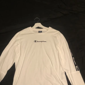 Vit långärmad Champion tröja - Säljer en vit långärmad tröja från Champion med svart logga på bröstet och Champion-tryck längs axlar och ärmar. Tröjan är oanvänd i stolen XS men passar även S.