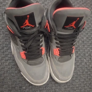 Nike Air Jordan 4 grå/svart/röd - Nike Air Jordan 4 sneakers i grått med svarta och röde detaljer. Skorna har meshpaneler, snörning och en synlig Air-sula. Insidan är fodrad med orange tyg och Jumpman-loggan syns både på plösen och hälen. Perfekt för dig som gillar klassiska och sportiga sneakers.