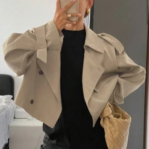 Beige croppad trenchcoat - Säljer en beige croppad trenchcoat med breda axlar och snygga detaljer som knytband vid ärmen och stora knappar framtill. Jackan har en loose passform och är perfekt för dig som gillar trendiga och stilrena plagg.