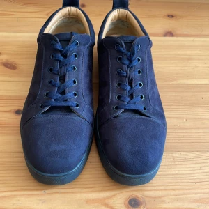 Mörkblå mockasneakers i Louboutin-stil - Snygga mörkblå sneakers i Christian Louboutin- stil i mjuk mocka med klassisk röd sula och rund tå i gott skick. Dustbag, skokartong och kvitto medföljer. Jag köpte dem här på Plick som äkta Loubotin-skor men när jag fick dem framgick det att skorna är välgjorda kopior. Därav ett lågt pris för ett par superfina skor. Skorna har snörning och diskreta detaljer, perfekt för dig som gillar lyxiga och stilrena sneakers med en twist.