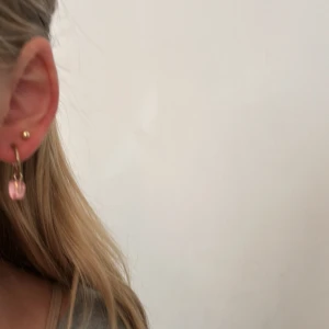 Guldiga örhängen med rosa hjärtan - Superfina örhängen i guldfärgad metall med hängande rosa hjärtan! Perfekt för dig som vill ha en söt och lekfull detalj till din look. Kommer aldrig till användning längre. Ignorera hur rött mitt öra är!!!😅 OBS, katt finns i hemmet!