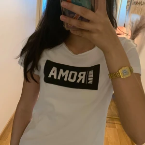 Vit Puma ROMA t-shirt i bomull - Vit t-shirt från Puma med svart tryck där det står ROMA på bröstet. Klassisk rund hals och korta ärmar. Materialet är mjuk bomull och passformen är normal. Perfekt för dig som gillar sportiga och cleana vibes.