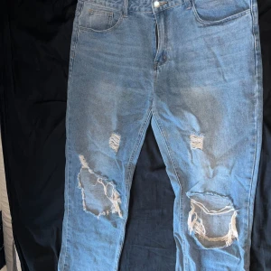 Ljusblå ripped jeans med raka ben - Säljer ett par ljusblå jeans med stora slitningar och hål på båda benen, raka ben och normal passform. Perfekta för en avslappnad streetstil och tillverkade i bomullsdenim.