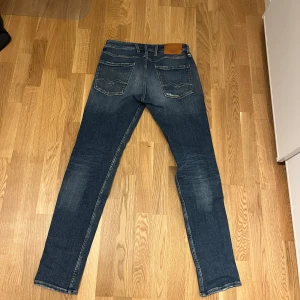 Replay Anbass jeans mörkblå herr - Snygga Replay Anbass jeans i mörkblå tvätt med slitningar och ljusare partier på låren. Klassisk femficksmodell med raka ben och normal passform. Jeansen har detaljerade bakfickor och läderpatch i midjan. Perfekta för en avslappnad och trendig look.