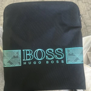 Svart axelväska från Hugo Boss - Snygg svart axelväska från Hugo Boss med justerbar rem och dragkedja upptill. Framsidan har ett turkost Boss-tryck och grafiska detaljer. Väskan är tillverkad i slitstark polyester och har en stilren, rektangulär form som passar perfekt för dina essentials.