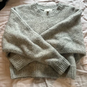 Grå stickad tröja med rund hals - Mysig grå stickad tröja med rundad hals och ribbade muddar vid ärmslut och nederkant. Perfekt för kyliga dagar och enkel att matcha med jeans eller kjol. Tröjan har en klassisk, loose passform och långa ärmar som ger en avslappnad vibe.