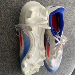 Adidas F50 fotbollsskor blå/röd - Säljer ett par Adidas F50 fotbollsskor med snygg design i vitt, blått och rött. Skorna har snörning, platt sula med dobbar och en sportig look. Ovandelen är i syntetmaterial och insidan är fodrad med blå textil. Perfekt för dig som vill sticka ut på planen.