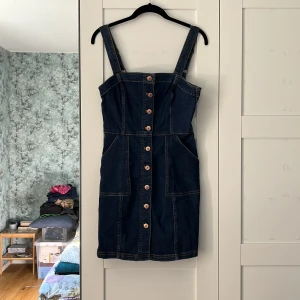 Jeansklänning H&M Divided - Mörkblå stretchig jeansklänning från H&M. Den har knappar på framsidan som går hela vägen, två justerbara axelband och två ganska djupa fickor i fram. Storlek: 36. Material: 82% bomull, 17% polyester och 1% elastan. Använd, men i väldigt fint skick!