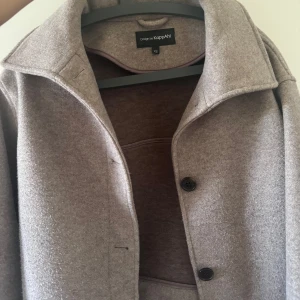 Beige kappa från KappAhl XS - Snygg beige kappa från KappAhl i storlek XS. Jackan har klassisk krage, två svarta knappar framtill och är tillverkad i mjukt ullmaterial. Den är croppad i modellen och har långärmade ärmar. Perfekt till hösten,  Lite nopprig 🍁