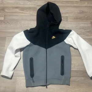 Nike hoodie med dragkedja och huva - Nike hoodie med cool färgblock-design i svart, grått och vitt. Tröjan har huva, hel dragkedja och två fickor med dragkedja framtill. Långärmad och med gul Nike-logga på bröstet. Perfekt för chill eller streetwear.