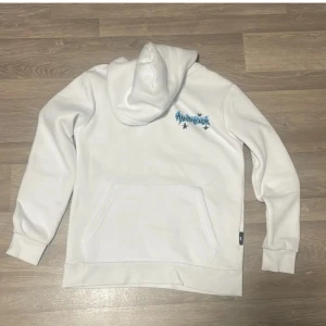 Vit hoodie från Hoodrich med blått tryck - Säljer en vit hoodie från Hoodrich med flera coola blå och svarta graffiti-tryck på ryggen och ett mindre tryck fram. Tröjan har huva, magficka och långa ärmar. Perfekt för dig som gillar streetwear och vill sticka ut med snygga detaljer.