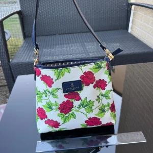 Ny Blommig axelväska  - Helt ny Snygg axelväska med vit botten och röda blommor samt gröna blad. Väskan har marinblå detaljer och justerbar axelrem. Insidan är randig i vitt och marinblått med dragkedjefack. Finns på Hägerstensåsen i Stockholm. 