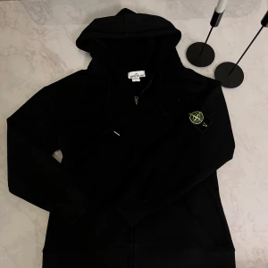 Svart hoodie från Stone Island - Svart hoodie från Stone Island med ikonisk patch på ärmen. Tröjan har huva med snörning, dragkedja framtill och långa ärmar. Perfekt för dig som gillar streetwear och vill ha en clean look. Dessutom går det att diskutera priset vid intresse!