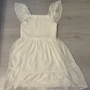 Vit kort klänning från Kappahl - Supersöt vit klänning från Kappahl med smockad överdel och volangig kjol. Klänningen har transparenta axelband med broderade blommor och är tillverkad i ett lätt, luftigt material. Perfekt för sommarens alla tillfällen!