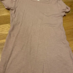 Beige kortärmad ribbad T-shirt - Säljer en enkel och stilren beige klänning med korta ärmar och rund hals. Klänningen är ribbad och har en rak passform, längden är cirka 57 cm och bredden 34 cm. Perfekt för dig som gillar minimalistisk och comfy stil. Skriv om ni har några funderingar!