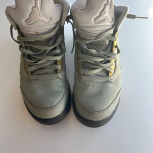 Nike Air Jordan 5 Retro Jade Horizon - Nike Air Jordan 5 Retro i färgen Jade Horizon/Desert Sand. Snygga sneakers med högt skaft, snörning och detaljer i ljusgrön och beige mocka samt svart sula med vit och beige mönstrad kant. Ikonisk Jumpman-logga på plösen och meshpaneler på sidorna.