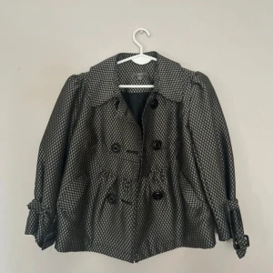 Mönstrad trenchcoat från apt.9 - Snygg trenchcoat från apt.9 i svart och silver med ett coolt rutigt mönster. Jackan har dubbelknäppning, bred krage och knytband vid ärmsluten. Passformen är loose och materialet känns som polyester. Perfekt för dig som vill sticka ut med din stil.