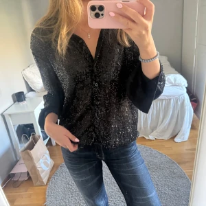 Svart paljettblus med knappar - Snygg svart skjorta med glittriga paljetter över hela tyget. Modellen har lång ärm och klassisk krage, samt knäppning framtill.🖤