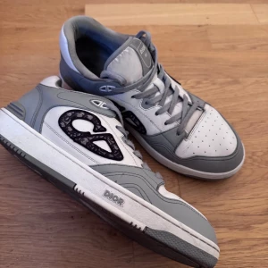 Dior B27 sneakers i grått och vitt - Säljer ett par Dior B27 sneakers i grått och vitt med coola svarta Dior-loggor på sidorna. Skorna har snörning, perforerad tå och en chunky sula med Dior-detalj. Materialet är skinn och textil, och designen är riktigt clean med en modern vibe. Äkta!!