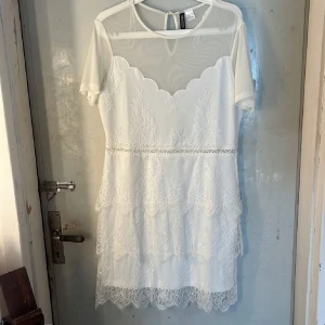 Vit spetsklänning med meshdetaljer - Supersöt vit kortklänning från Chicoree med genomskinlig mesh upptill och vackra spetsdetaljer över hela klänningen. Klänningen har korta ärmar, rund hals och ett romantiskt spetsmönster med volanger nedtill. Perfekt för student eller sommarens alla tillfällen!