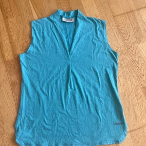 Turkos ärmlös linnetopp från SKHOOP - Snygg turkos linnetopp från SKHOOP med v-ringad hals och liten krage. Toppen är ärmlös och har en enkel, clean design som passar perfekt till jeans eller shorts. Mjuk och lätt bomullskänsla, perfekt för varma dagar. Toppen är aldrig använd!