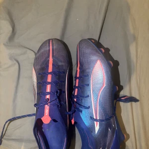 Blå fotbollsskor Puma Ultra 41 - Säljer ett par Puma Ultra fotbollsskor i storlek 41. Skorna är blå med rosa och vita detaljer, har snörning och en platt sula med dobbar för gräsplan. Ovandelen är tillverkad i syntetmaterial med ett sportigt mönster och smal passform. Perfekt för dig som vill ha speed på planen.