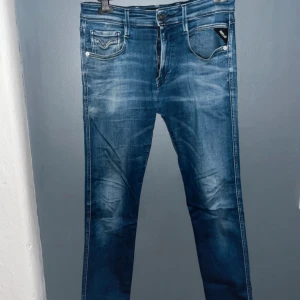 Replay Anbass blå jeans herr - Snygga Replay Anbass jeans (32x36) i klassisk blå tvätt med slitningar och kontrastsömmar. Modellen har fem fickor, dragkedjegylf och smal passform. Perfekta för dig som gillar en modern och stilren look. Jeansmaterialet är mjukt och följsamt. 
