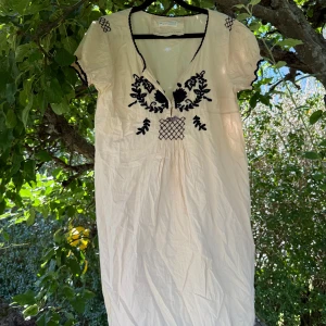 Odd Molly ljus kortklänning med broderi - Superfin beige kortklänning från Odd Molly med svart broderad detalj framtill och på axlarna. Klänningen har v-ringning, korta puffärmar med vågig kant och ett sött litet rosettband. Perfekt för sommarens alla tillfällen! Storlek 3 som enligt deras storleksguide motsvarar en storlek L.  Mycket fint skick, jag hittar inga synliga anmärkningar eller defekter. 