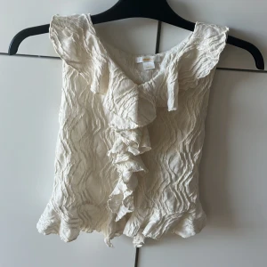 Vit volangblus från Gina Tricot - Supersöt vit blus från Gina Tricot storlek 156/164 med volanger längs halsringningen och nederkanten. Blusen är ärmlös och har ett crinklat, har använt 2 gånger. Ingen fläck eller skada, 