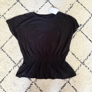 Svart topp med paljetter från H&M - Svart topp från H&M med coola paljettdetaljer framtill. Modellen har korta ärmar och en smockad midja som ger en snygg form. Passar även S/Xs.