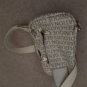 Beige axelväska med textmönster - Snygg beige axelväska från Generation Unique med tryckt textmönster över hela väskan. Väskan har justerbar axelrem, två fack med dragkedja i guld och detaljer i syntetmaterial. Perfekt för att bära dina essentials och ge din outfit en cool touch.