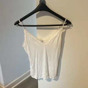 Vit ribbad linnetopp med spets från MISU - Supersöt vit linnetopp  med tunna axelband och ribbad struktur. V-ringad fram och bak med fina spetsdetaljer längs kanterna som ger en romantisk vibe. Perfekt att styla med jeans eller kjol för en chill look.