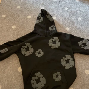 Svart hoodie med grått mönster - Säljer en svart Denim Tears hoodie med huva och stora gråa blommönster över hela tröjan. Hoodien har en klassisk känguruficka framtill och långa ärmar. Perfekt för dig som vill vara lite extra premium. 100% äkta