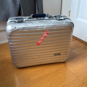 Rimowa bag - Säljer en stilren och robust resväska från Rimowa i silverfärgad aluminium. Insidan är blå med mönster och har justerbara remmar för att hålla packningen på plats. Ena spännet knäpper inte lika bra som det andra men fullt funktionabel. Nycklar finns ej