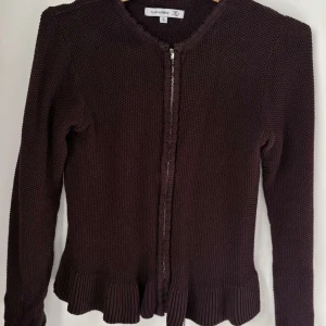 Stickad kofta från Flash Woman - Säljer en stickad kofta från Flash Woman i storlek S. Koftan har långärmade ärmar, dragkedja framtill och en snygg ribbad volangkant nedtill som ger extra stil. Perfekt för dig som gillar klassiska detaljer och vill ha något mysigt till hösten.