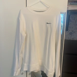 Vit Nike sweatshirt med logga - Snygg vit sweatshirt från Nike med klassisk svart swoosh-logga på bröstet. Tröjan har rund halsringning och långa ärmar. Perfekt för en clean och sportig look. Tillverkad i mjukt material som känns skönt mot huden.