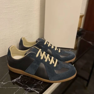 Maison Margiela blå sneakers - Maison Margiela sneakers i mörkblå mocka och skinn med beige snörning och innersida. Snygg kontrast mellan material och färger, samt klassisk gummisula med logotyp under. Kvitto och box finns.