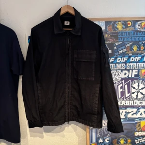 Svart vindjacka från C.P. Company - Snygg svart vindjacka från C.P. Company med diskret meshfoder och praktisk dragkedja framtill. Jackan har en cool, urban look med märkets logga på insidan och på dragkedjans band. Perfekt för dig som gillar stilrena och funktionella plagg.