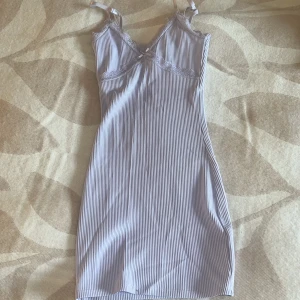 Ljuslila ribbad kortklänning med spets - Supersöt kortklänning med tunna axelband, ribbad struktur och spetsdetaljer vid v-ringningen. Liten rosett framtill ger extra gullig vibe. Perfekt för dig som gillar en feminin och enkel stil.