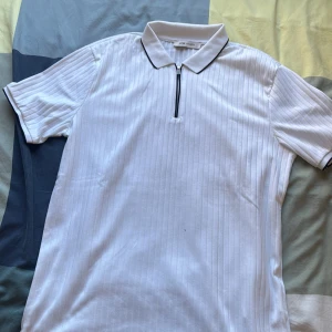 Vit ribbad t-shirt med dragkedja - Snygg vit t-shirt från Low Vision med diskreta vertikala ränder och ribbad struktur. Har en klassisk krage med svart kant och en dragkedja framtill. Perfekt för dig som gillar stilrena detaljer och sportig vibe.