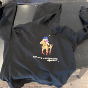 Ralph Lauren Hoodie - Svart hoodie från Polo Ralph Lauren i storlek XL med klassisk broderad Polo Bear på bröstet. Tröjan har huva och långa ärmar, tillverkad i mjuk bomull. Snyggt tryck med färgglada detaljer som gör den unik och stilren. Är XL men sitter som en small på mig och jag är 174. Pris ej hugget i sten.